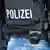 Der Rücken eines bewaffneten Polizisten, auf dessen dunkelblauer Schutzweste in weißer Schrift "Polizei" steht 