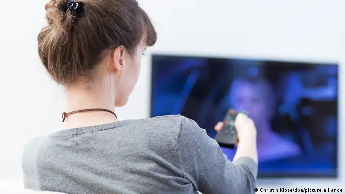 A young woman shown from behind, pointing a remote control with her right hand towards a TV screen.