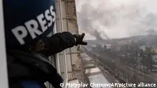 Someone wearing a press helmet points to smoke rising from a building far away