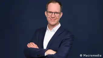 Florian Meißner is professor for media management and journalism at Macromedia, University of Applied Sciences in Cologne, Germany. He is wearing a dark suit with a white shirt, and his arms are crossed in a comfortable possition. He is wearing glasses and he is smiling. 