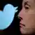 On the left half of the picture, slightly blurred in the background, the Twitter logo and in the right half of the picture, in focus in the foreground, Elon Musk with a sceptical expression on his face.
