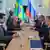 Pessoas reunidas em torno de uma mesa. Ao fundo, uma bandeira do Brasil e uma da Rússia