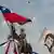 Homem empunha bandeira chilena em cima de uma estátua. Ao lado, uma imagem de Víctor Jara.