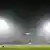A cricket pitch lit up at night. The stands in the background are barely visible through the smog