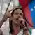 Maria Corina Machado fala ao microfone, com uma bandeira da Venezuela ao fundo