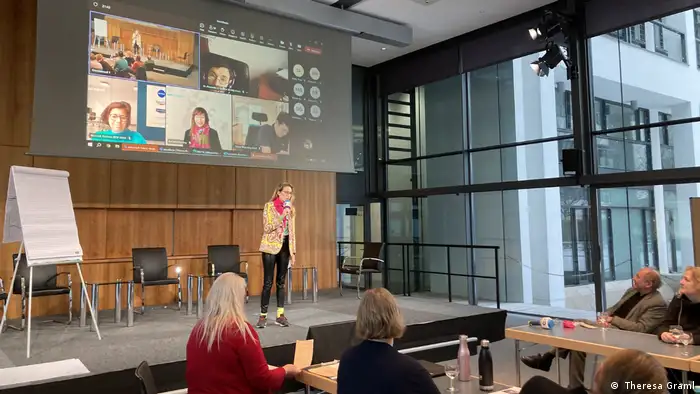  The woman on the stage, Zahra Nedjabat, speaks into a microphone in front of a group of listeners. On a projector screen behind her, people are taking part in the meeting via Teams video link.