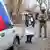 Сотрудница избирательной комиссии держит урну во время досрочного голосования, за ее спиной стоит военный с автоматом, Донецк, 14 марта 2024 года